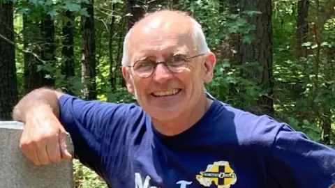 Sean Drysdale who is bald on top and has grey hair at the side and is wearing a Norn Iron blue T-shirt and grey shorts stands beside a a stone sign that says Old Belfast with a forest in the background