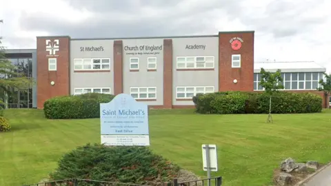 Street view image of St Michael's Church of England High School, a rectangular two-storey grey rendered building with red brick pillars either side and white windows behind a large lawned area to the front.