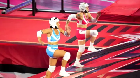 BBC/Hungry Bear/David MacCormack Rachael Phillips with red and white sports top and bottoms, white helmet, white trainers and tattoos on her arms. Naomi Church with blue and white sports top and bottoms, white helmet and white trainers. They are running up the red, white and black travellator. 