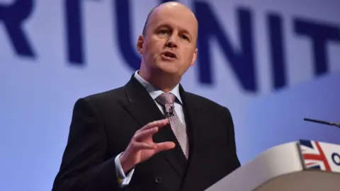 BBC A photo taken at a conference with Sir Ashley fox wearing a suit, speaking at a lectern with one of his arms raised slightly. 