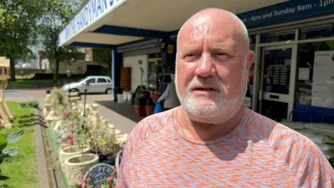 Rob Evans is pictured outside a retail premises. He is bald with a white beard and is wearing an orange and grey patterned t-shirt.