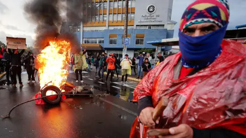 EPA In the background a group gathers around a tire fire in the middle of a street, near a government building while in the foreground a man in a red poncho, blue balaklava and patterned red hat stares out at the camera 