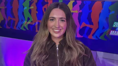 Molly, who is wearing a brown jacket, is sitting in the BBC Radio Manchester studio. There is a BBC-branded purple microphone on a desk in front of her. 