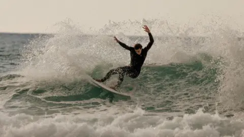 @bellarosebunce A surfer is riding across the face of a breaking wave on a surfboard, lifting the board up the lip while water sprays behind. Another figure is visible in the water nearby as the wave rolls in under a grey sky.
