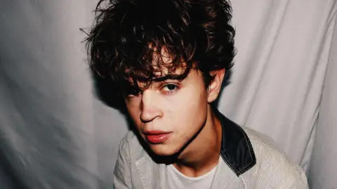 Tom A. Smith has dark curly hair which slightly covers his eyes. He is wearing a light-coloured jacket with darker vertical stripes and a dark colour. He is posing against a white sheet background
