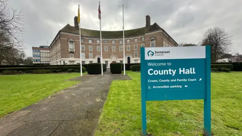 A turquoise sign reading welcome to county hall standing in the grass outside a curved stone municipal building with three flags flying against a grey sky