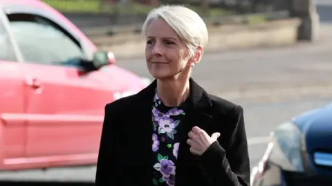 PA Media Lady Eleanor Donaldson has short blonde hair. She is wearing a black blazer and a floral blouse. Behind her a red car can be seen.