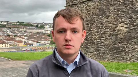 Pádraig Delargy has short brown hair. He is is wearing a grey fleece and a blue shirt. He is standing near to Derry's historic walls and a number of houses in the Bogside can be seen in the background.