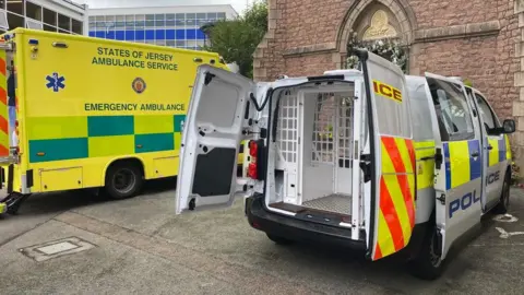 A white police custody van is parked outside a school with the back doors open showing a seating area at the back with bars around it. A yellow ambulance is parked nearby and the open back door and ramp are just in shot.
