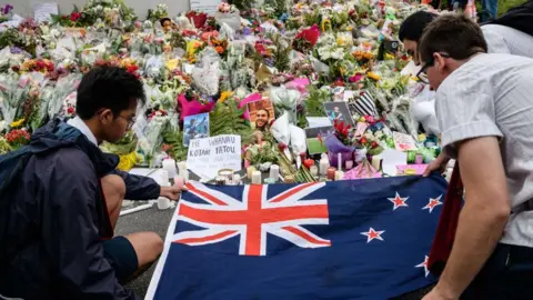 Three men lay the New Zealand flag over a spread of flower bouquets offered for victims of the 2019 Christchurch mosque shooting.