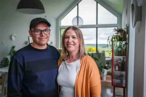 Chris Labrooy in a navy striped top and a black skip cap stands in his winning living room with wife Jessica Zanoni who is a blonde lady wearing a white vest and orange cardigan