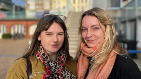 BBC / George Carden Two women - one with black hair and a leopard print scarf - and another - who is blond with a coral scarf - look at the camera with steps to the entrance of the i360 tower in the background