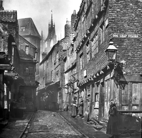 Newcastle Local Studies Library/Tyne Bridge A black and white view up a street of old buildings with women standing outside