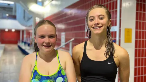 Eleanor and Parry are in in swimsuits stand on the poolside of an indoor swimming facility. The tiled pool deck extends into the background, with red tiled walls, pool signage and swimming lanes visible.