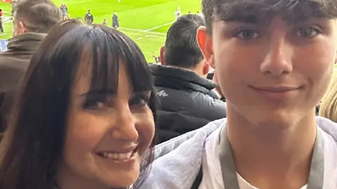 BBC presenter Lucy Owen on the left who is smiling next to her son Gabriel. He is wearing a white t-shirt and grey hoody. Behind them is the Etihad Stadium