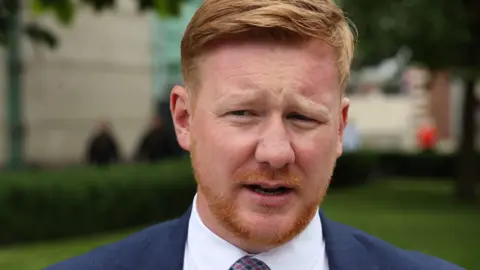 Daniel McCrossan speaking to people outside a building. He has short ginger hair and beard and is wearing a navy suit with a white shirt and a red patterned tie.
