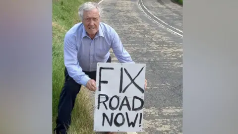 Redcar and Cleveland Councillor Steve Kay is standing at the side of a single carriageway road holding a homemade white sign with the words "Fix road now!" written in black. He has short white hair and is wearing a blue shirt and blue trousers.