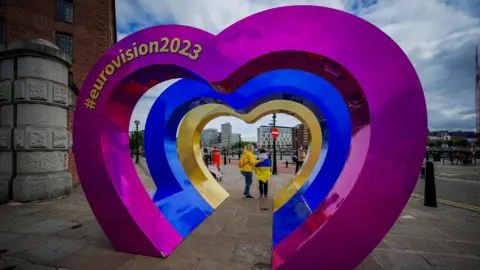 A series of heart shaped structures. One is pink, the second is blue and a third is golden. Eurovision 2023 is written on the pink heart structure in gold writing. Two people can be seen through the centre of the hearts. 