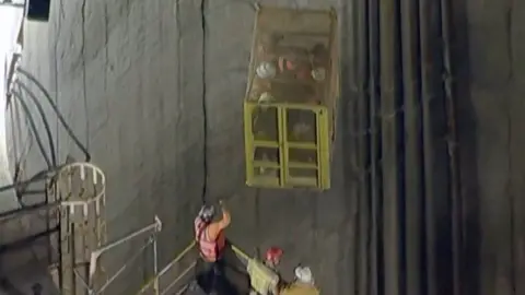People are in a cage being lifted up an industrial tunnel.