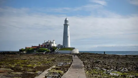 The concrete causeway leading to the lighthouse at St Mary's island on a sunny day. The causeway us surrounded by rocky terrain.