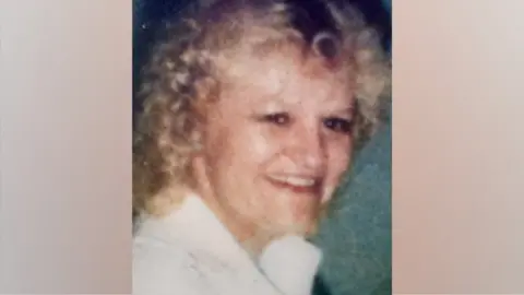 A picture of Patricia, who has light‑coloured, curly hair styled close to the head and is wearing a light‑coloured collared garment. The overall image quality shows visible grain and fading, with subdued colours and softened edges that indicate age or wear