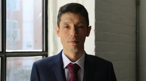 Person in a suit and red tie standing beside a large window in an office-like setting.