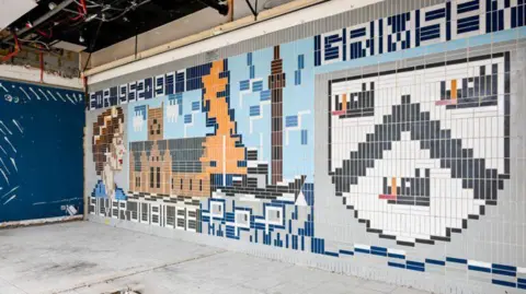 The tiles are thin and rectangular and are laid vertically.  There is an image within the tiles design of the Queen, a church, a map of Great Britain and a coat of arms.  Writing in the tiles says ER 1952.1977, Grimsby, Silver Jubilee.