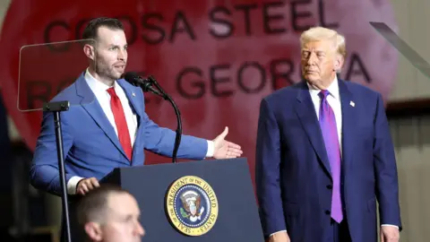 A Man in a navy suit and tie stands at a podium gesturing with his arm toward Donald Trump who is also wearing a navy suit