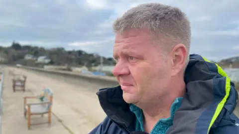 Jake Bowers looks out to sea. There are benches on the promenade next to him. The sky is cloudy and he is looking pensive.