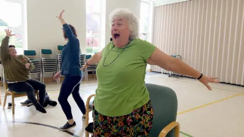 A woman in a green top and colourful trousers jumps out from her seat with her arms outstretched and a big grin on her face