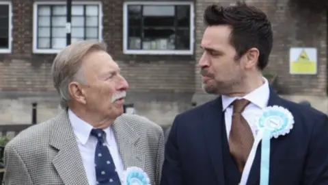 Alex Sibley A photo of two men wearing Reform Party rosettes, looking quizzically at each other outside an industrial brick building. The man on the left is older, with grey hair and a grey moustache, and wears a black and white houndstooth jacket over a white shirt and a navy tie. The man on the right has black hair in a duck tail style and wears a navy suit, a white shirt and a brown tie. 
