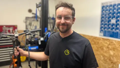 A man with brown hair and clear glasses on smiles directly into the camera, his right hand is holding the handles of an orange bike. Behind him are a wall with tools on in his workshop. He is wearing a black t-shirt with a bright yellow logo on the left side of his chest. 