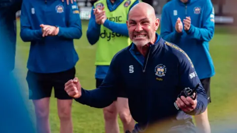 BUCS A close-up of a man on a rugby pitch looking ecstatic - crouching down and holding his fists up in triumph. There are three players behind him shown from the neck down, all clapping.