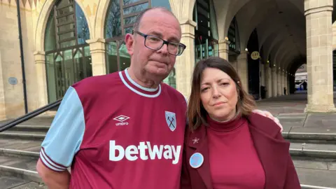 Kate Bradbrook/BBC Luke's parents Richard Abrahams and Julie Needham are looking at the camera. He is wearing a burgundy and blue football shirt, and has balding hair and spectacles. She is wearing a red pullover and burgundy jacket and has shoulder length brown hair. He has his arm around her shoulder and the pair are standing near some steps leading to an arched pedestrian walkway.