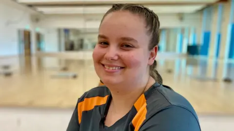 A young woman with her brown hair tied back in a french plait wears a green sports top with orange stripes on the shoulders and smiles in an exercise studio