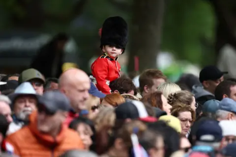 Reuters A small child wearing a bearskin hat and red uniform looks on as they sit on someone's shoulders with other members of the crowd next to them.
