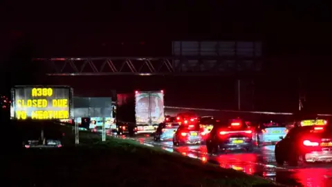 Michael Ormsby Standstill traffic on the A380. It is raining and car brake lights are glowing in the dark. A sign to the left of the road says 'A380 closed due to weather'.