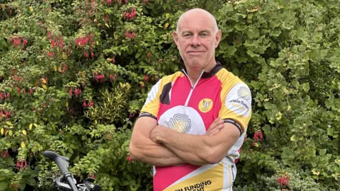 Brain Tumour Research John Starns wearing a pink, yellow and white cycle top looking to camera in front of a hedge
