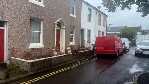A picture of Adam Carruthers childhood home which is a terraced house with pebbledash and a central red door. There is no pavement so from the door there is a small, walled space which steps straight on to the road. Two vans are parked outside and a silver Volvo is driving towards the camera through a narrow gap.