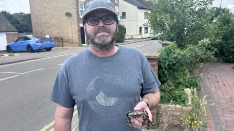 Kate Bradbrook/BBC A man in a light blue T-shirt, baseball cap and glasses standing in front of a residential road while holding a black and beige crab.