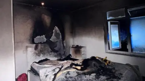 A fire-damaged room. There is dark scorching and smoke on the walls and the bedding is burnt. The windows are open and a fire hose can be seen balanced on the bed.