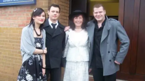 Family photograph A family photograph of  Mr Watson with his mum, sister and brother. They are dressed in smart clothes, standing outside a church. They are smiling at the camera and have their arms around each other. 