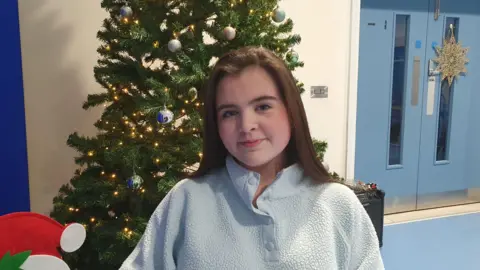 A young girl with long brown hair. She is wearing a light blue fluffy top and is standing in front of a decorated Christmas tree