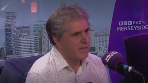 A middle aged man with grey hair, wearing a white shirt with a dotted pattern, sits in a radio studio speaking into a purple microphone. In the background is a photo of a city skyline and a purple logo reading BBC Radio Merseyside.
