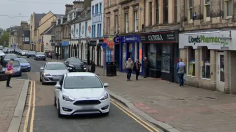 A google view of cars driving past pedestrians outside shops on Elgin High Street