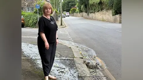 A woman wearing black trousers and a top standing on a pavement next to a street. The picture shows a damp area where water has been flowing. 