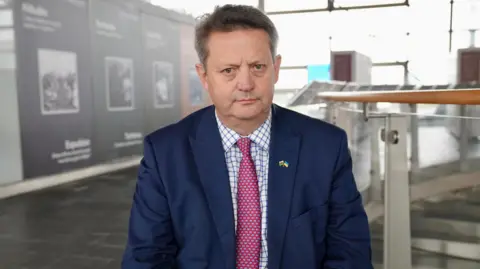 Alun Davies MS is wearing a suit and tie will posing for a photograph in the Senedd. He is wearing a royal blue suit with a white and blue checked shirt and a pink tie with a blue pattern on 