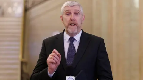 PA Media John O'Dowd speaking in Stormont. He is wearing a black suit, blue patterned tie and a white shirt. He has grey hair.