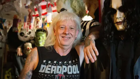 Martin, who has tattooed arms, stands in a room densely filled with collectables. A black-and-white striped character figure stands behind him, while another life-sized figure with long black hair stands beside them. Shelves and walls are covered with toys, posters, masks and decorative objects.