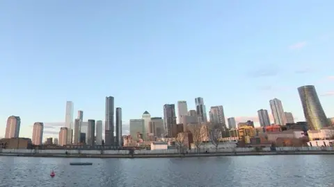 A body of water with tall commercial buildings in the background, two non-functioning cranes stand over the wharf